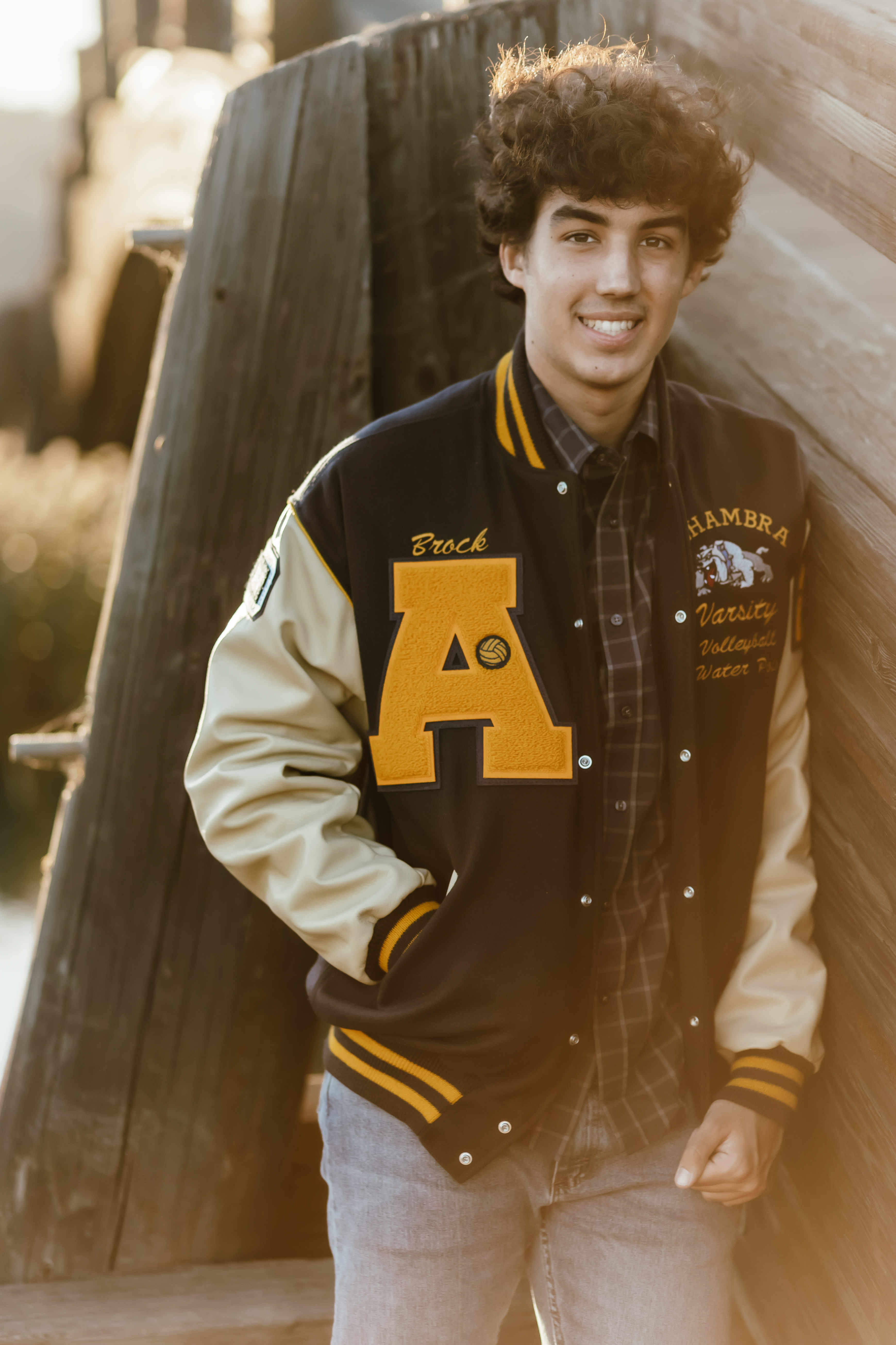Teen boy wearing letterman jacket leaning against wooden bridge.