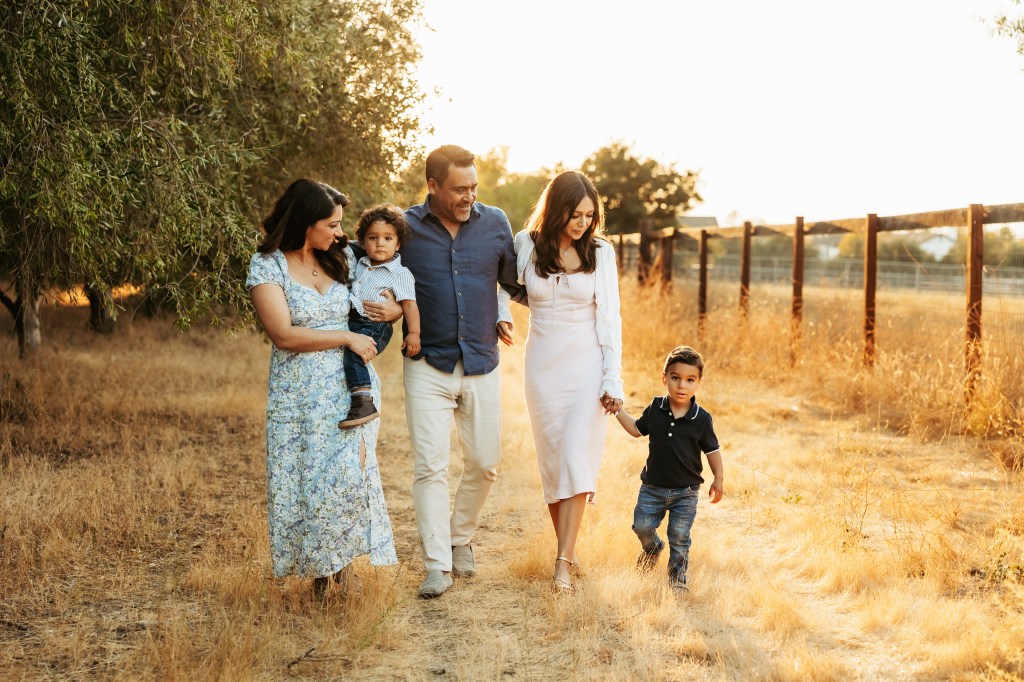 A family of five; mother, father, teen daughter and two toddler boys walk between olive trees and a fence bathed in golden light