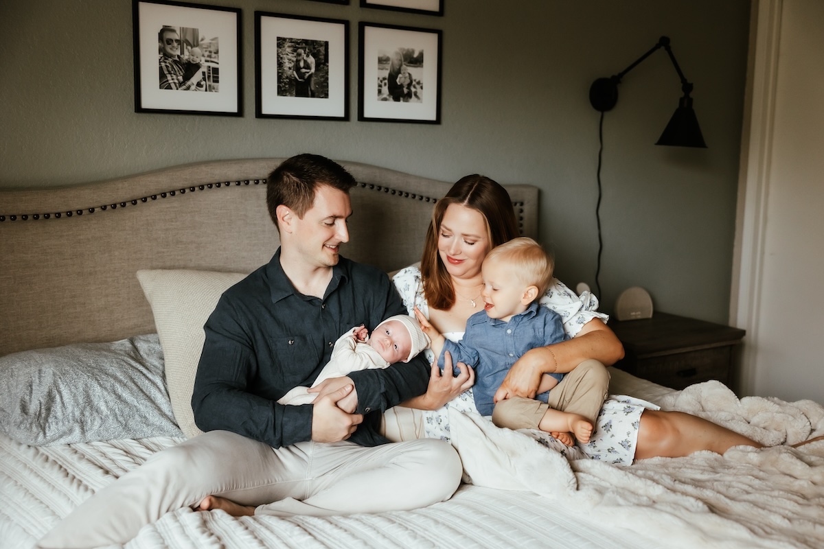 A family of four sit on a bed, toddler is patting the newborn baby's head.