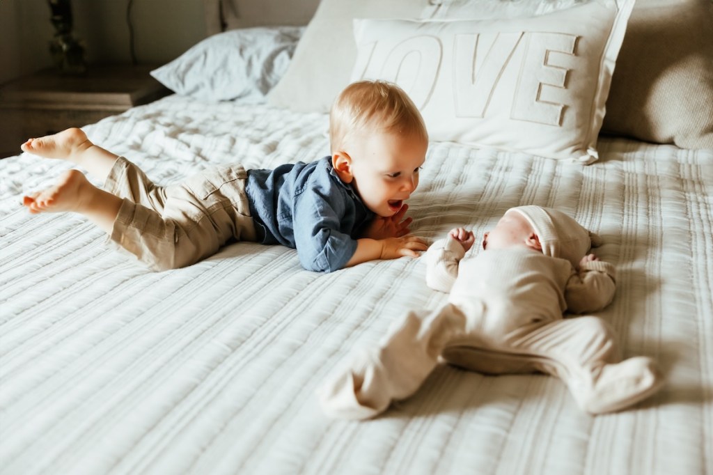 Toddler brother and newborn baby brother look at each other while laying on their parent's bed.