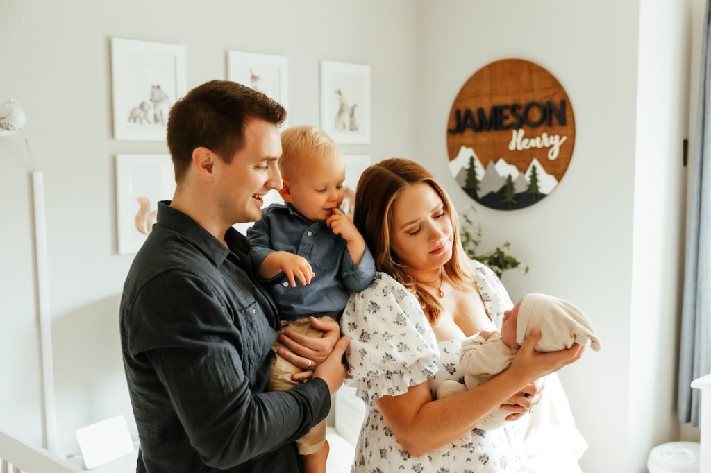 Mom hold up newborn baby brother while dad hold big brother toddler in the nursery for photos.