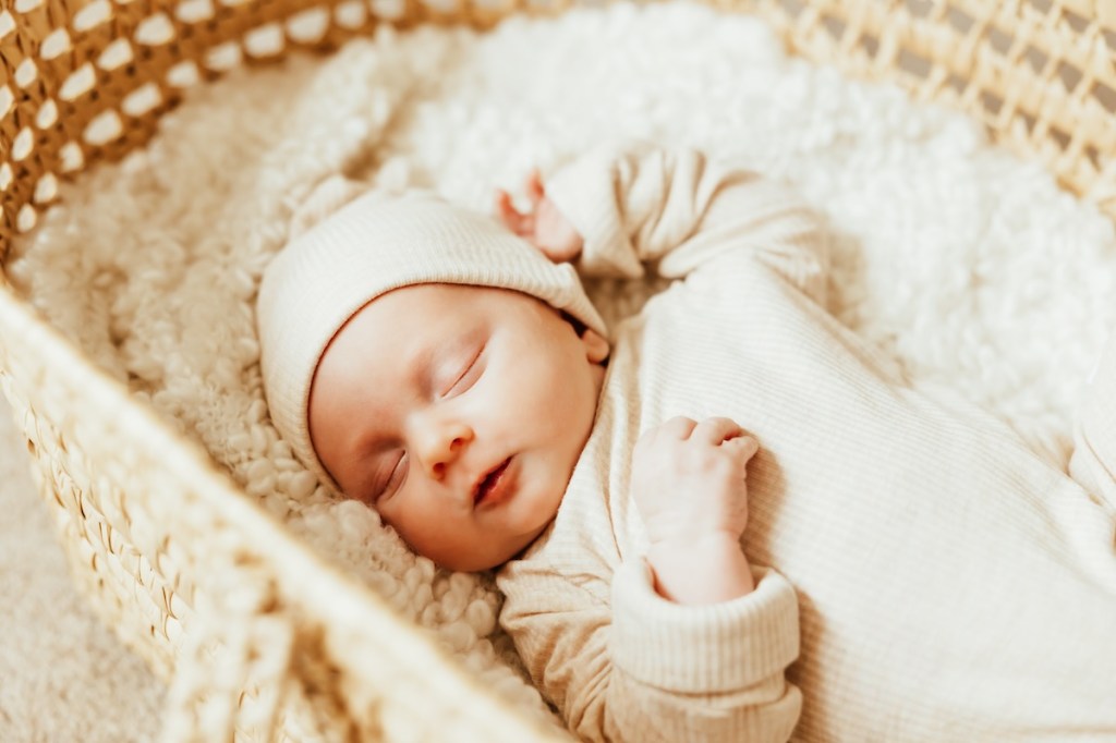 A sleeping baby wearing a cream colored sleeper set lays in a woven basket for newborn photos.