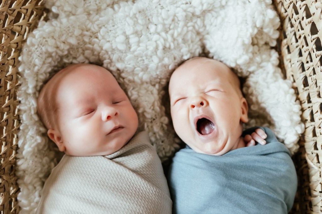 Twin babies lay side by side in a woven basket. They are swaddled and sleeping, on baby is yawning.