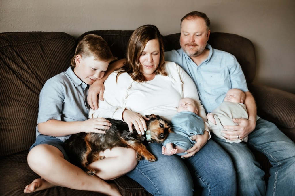 Mother, father and big brother sit on the couch together with 2 newborn babies and the family dog in their laps.