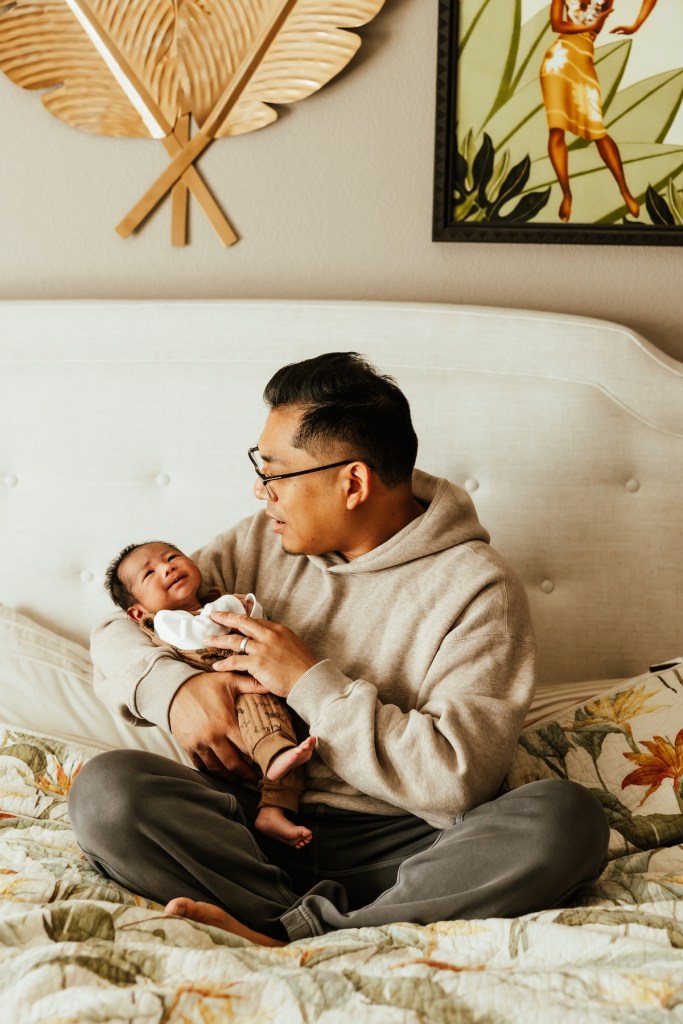Dad dressed in sweats cradles his newborn son in his arms while sitting a queen size bed.