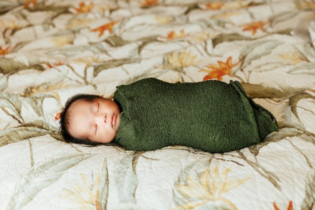A newborn baby swaddled in a forest green textured blanket lays on a tropical bedspread.