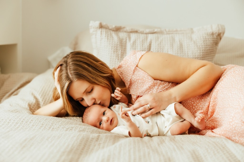 Mother kisses newborn baby's head while side lying on the bed.