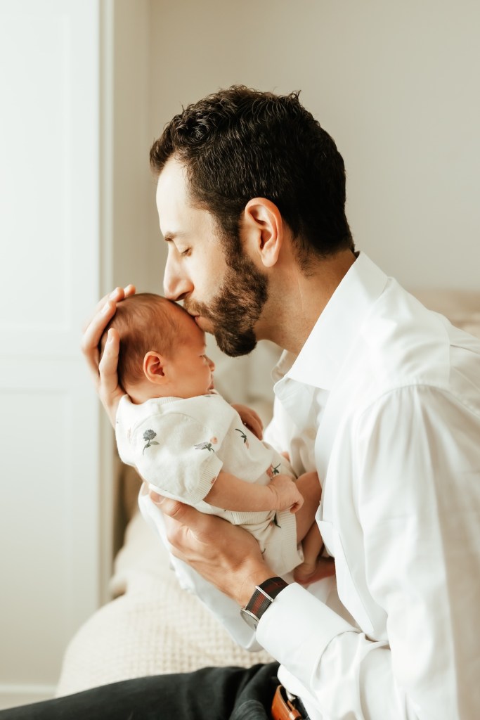 Dad kisses newborn baby's head lit by natural window light.