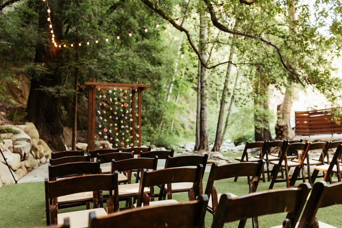 Ceremony site under the redwoods and beside a creek. Empty chairs and bistro lights lead to a wooden pergola decorated with hanging flowers by Zaddy Blooms. Redwood Grove in Saratoga Springs, California.
