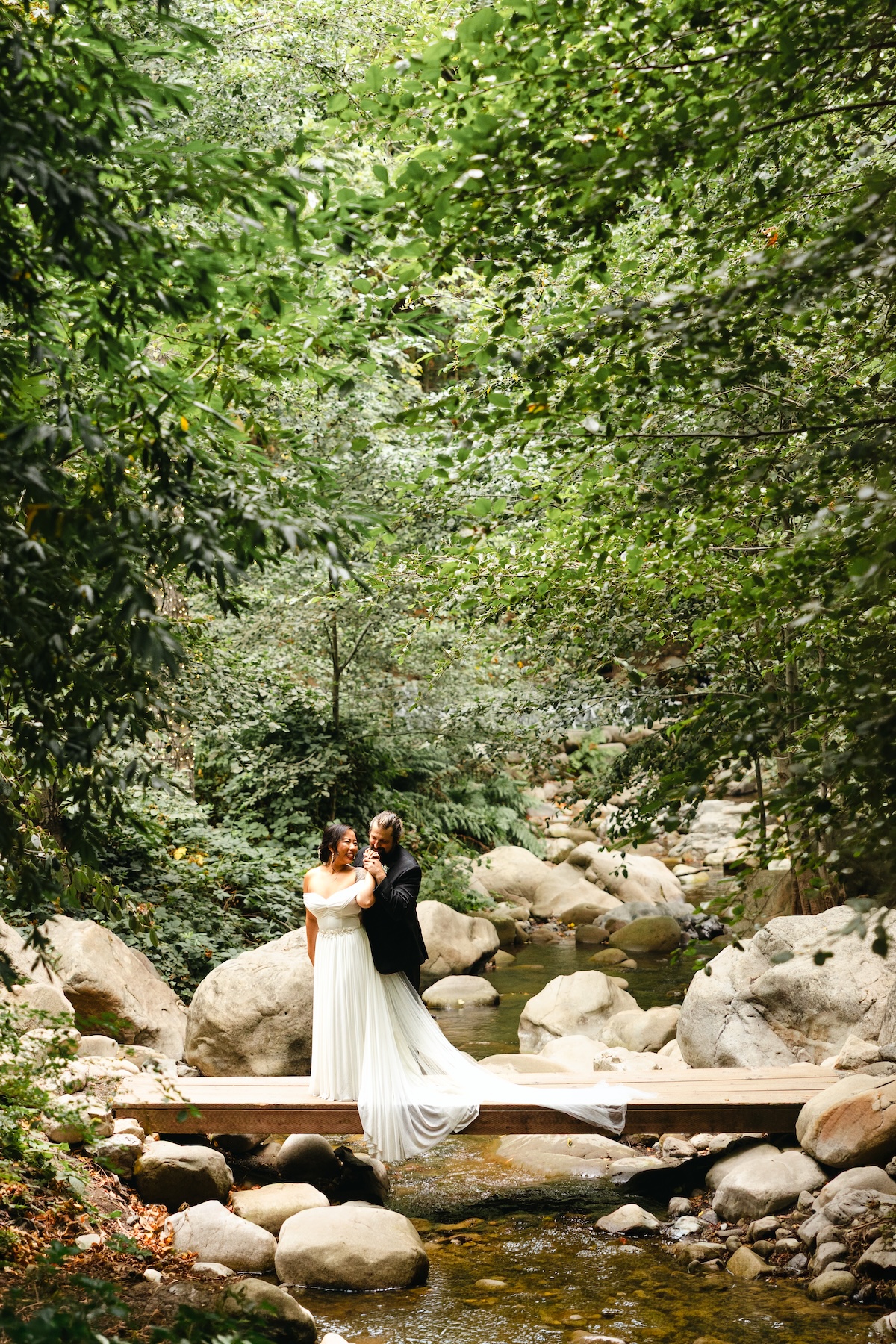 Bride and Groom stand on a small wooden bridge over a creek in a forest wedding venue, Saratoga Springs California.